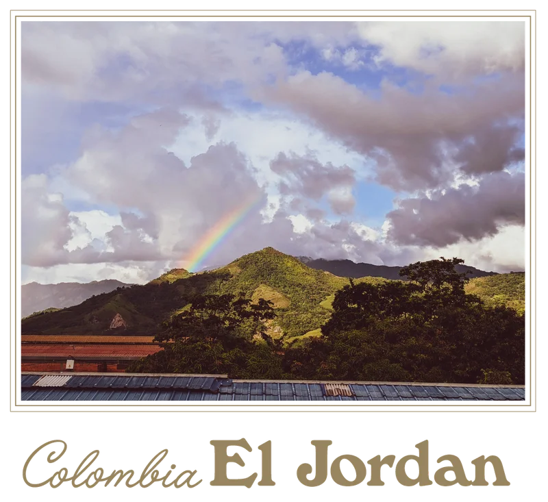 A rainbow arcs over the lush green hills of El Jordan coffee farm in Colombia, with dramatic clouds overhead and treetops and rooftops in the foreground.