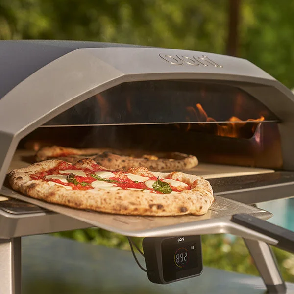 A person taking a pizza out of a Koda 2 Max oven, while another pizza cooks inside simultaneously
