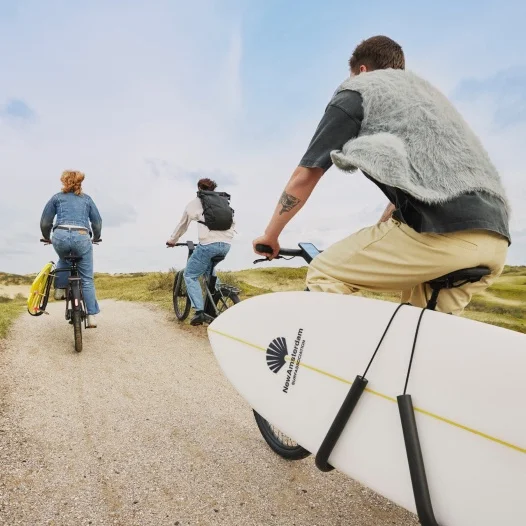 Three people cycling on Cowboy bikes along a sandy trail. The rider in the foreground is carrying a surfboard strapped to the side of their bike. The background shows grassy dunes and a bright sky.