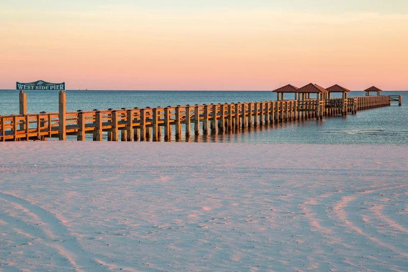 A far-reaching pier with small canopies along the way on a wide beach at sunset.