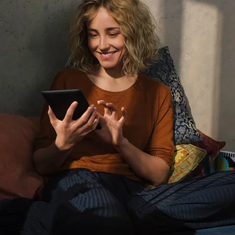 A smiling young woman sitting on bed using E-book reader as sunlight from a window casts a column of light from her face down to her hands holding the e-reader.