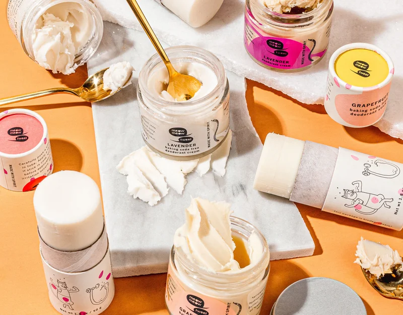A collection of deodorant cream in jars and deodorant sticks in paper tubes lay on an oragne backdrop with slabs of marble and spoons