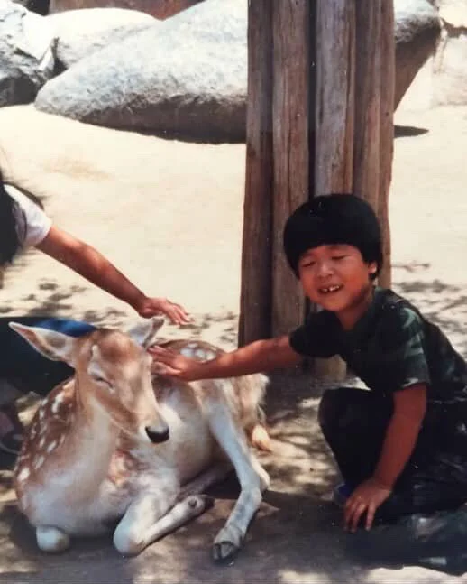 A child crouches down to pet a deer and smiles at the camera with their eyes closed.