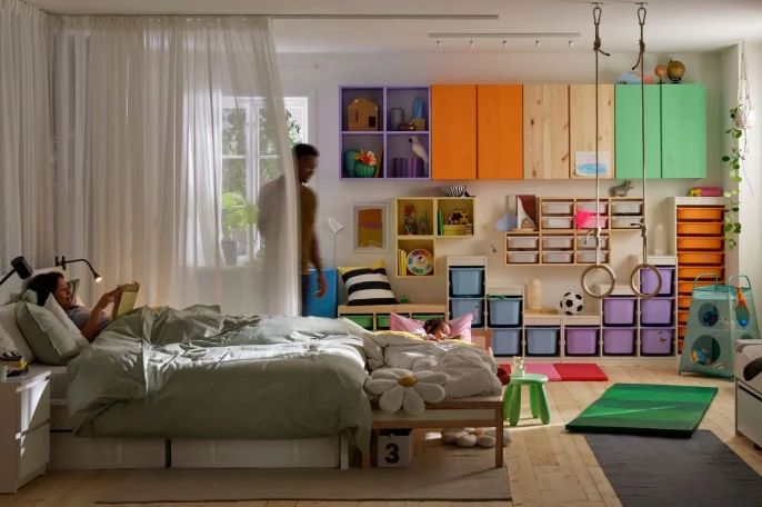 A room with two adults and a child. The child and one adult lie in separate beds. The other adult is standing by a curtain.