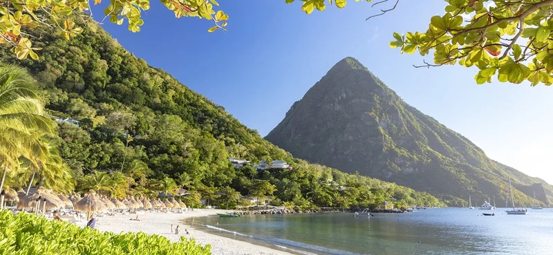 A pristine beach with white sand, lush green hills, and a towering mountain in the background, framed by tropical leaves