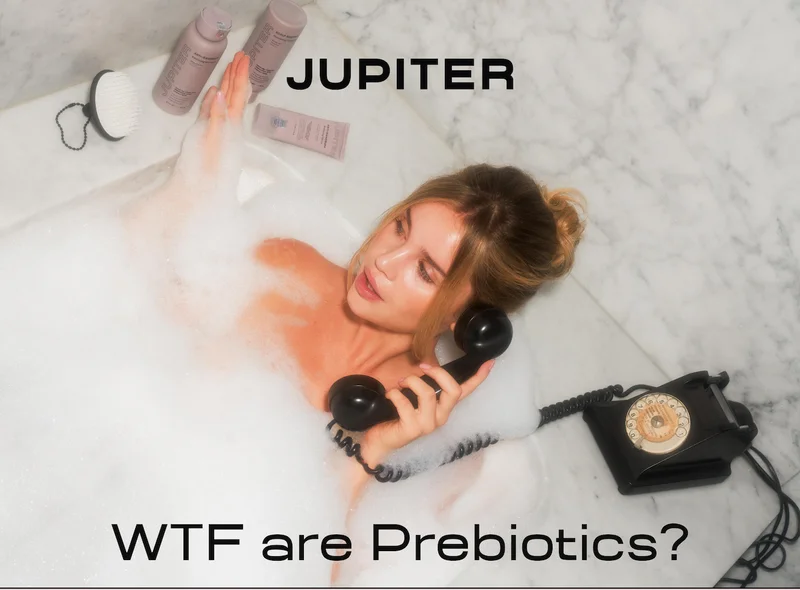 Woman relaxing in a bubble bath holding a vintage rotary phone receiver, with Jupiter Anti-Dandruff Balancing Shampoo, Scalp Soothing Conditioner, Anti-Dandruff Restoring Serum, and a Jupiter scalp brush arranged on the tub edge. Headline reads "WTF are Prebiotics?"