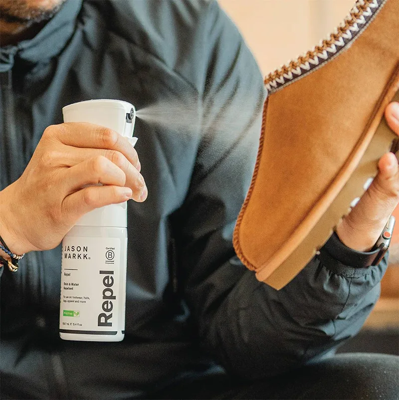 A medium shot shows a person holding a white spray bottle labeled 'JASON MARKK. Repel Stain & Water Repellent' and spraying it onto a tan suede slippe...