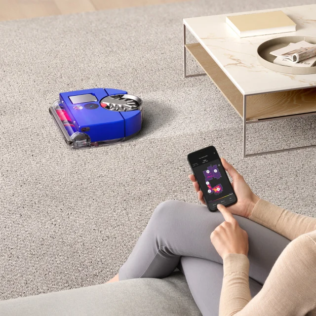 A Dyson 360 Vis Nav robot vacuum cleans while the owner types settings in the mobile app