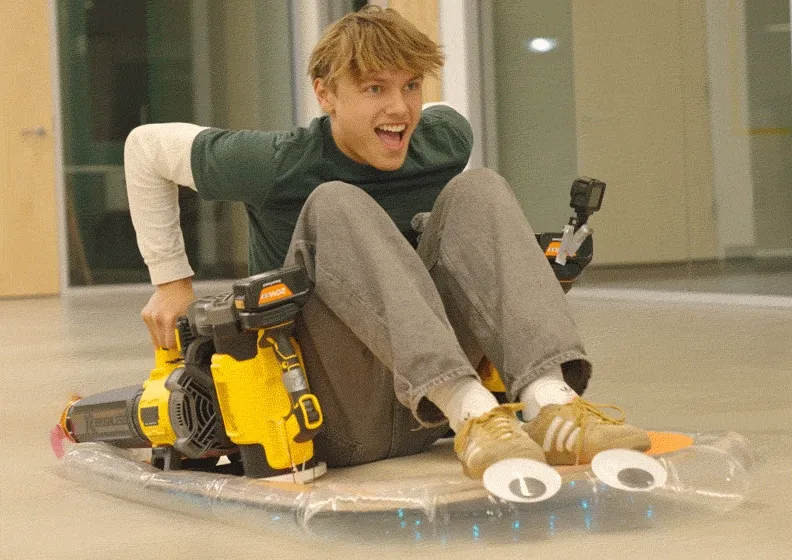 A teen rides a homemade hovercraft powered by a leaf blower across a smooth floor.
