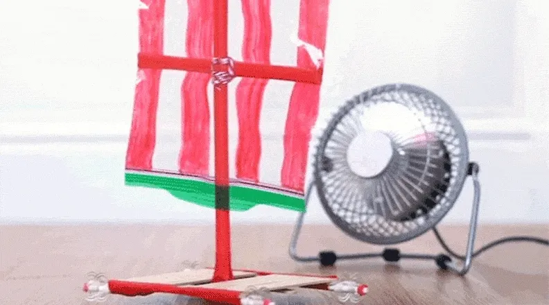 A red-and-green DIY wind car zooms forward as a fan blows air toward its sails.