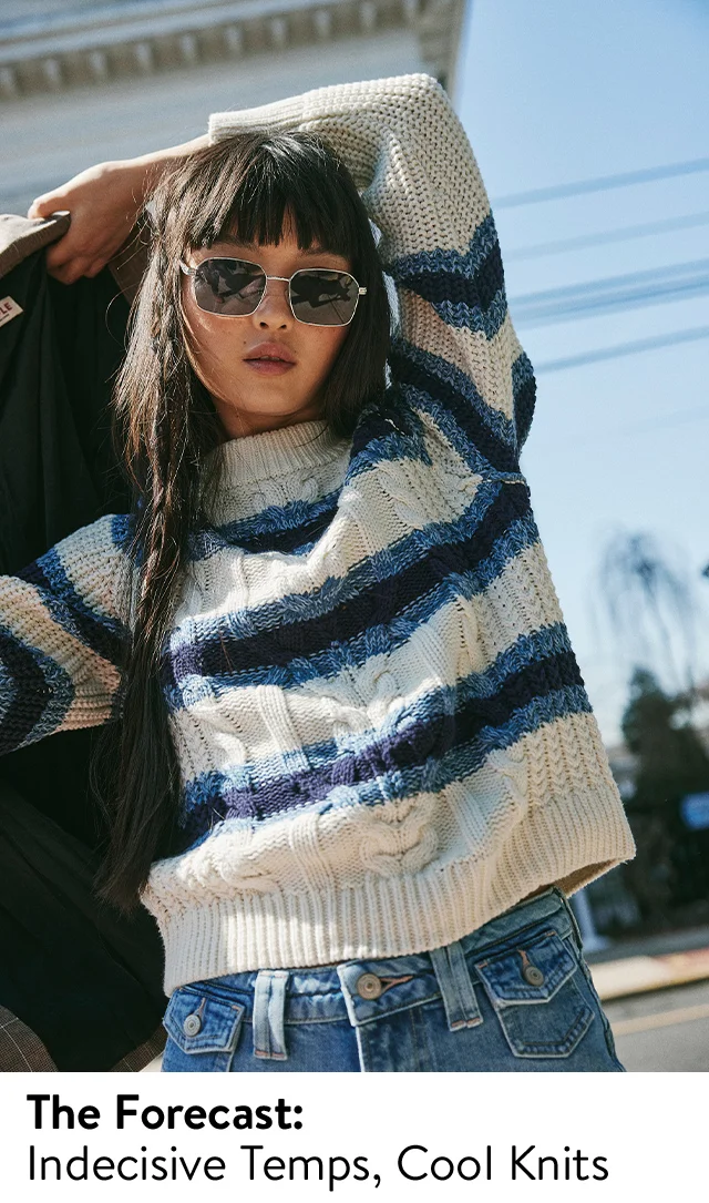 A woman wearing a striped cable-knit sweater, sunglasses and jeans.