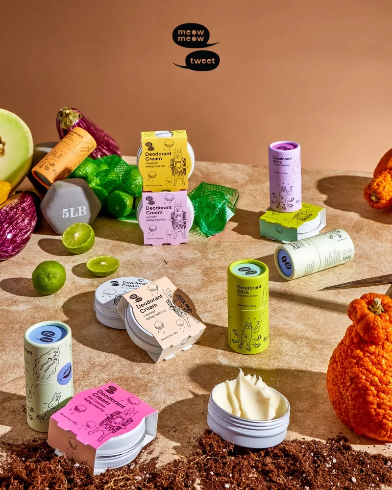 Many colorful tins and sticks of deodorant are arranged on a brown marble counter top with various fruits and veggies.