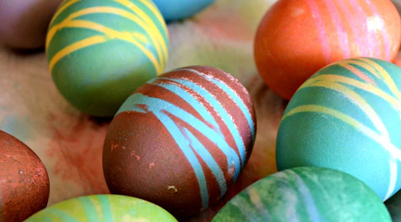 A collection of dyed Easter eggs with intricate patterns created by wrapping rubber bands around them before dyeing. The text reads, ''Rubber Band Eggs – Make beautiful designs with this egg-dyeing technique.''