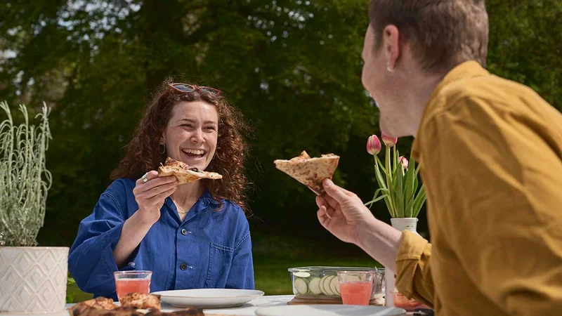 A man and woman sharing a pizza, on a bench in a garden