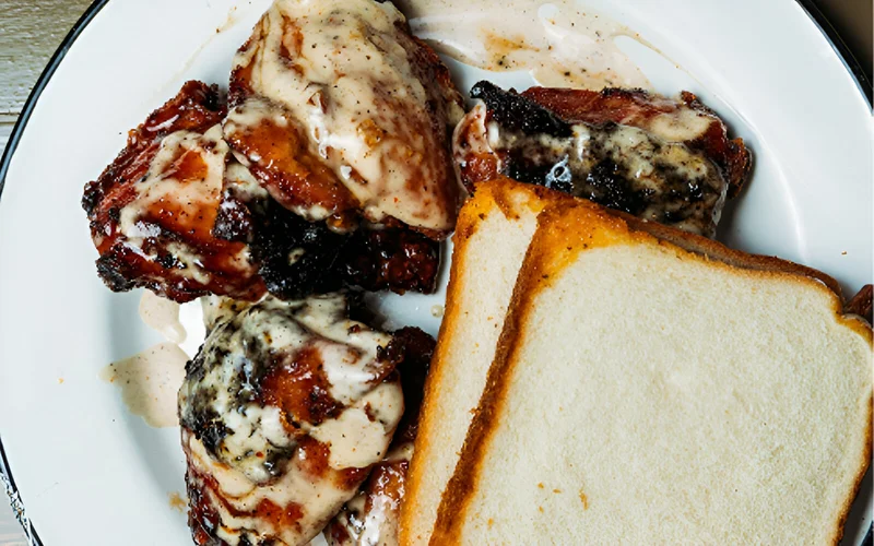 A plate of four pieces of BBQ chicken and two slices of white bread, covered in white sauce.