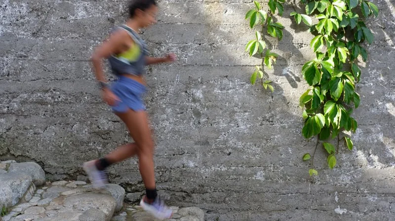 The blurry form of a runner wearing blue shorts and a yellow vest moves down stone steps. A green vine hangs from a stone wall next to her.