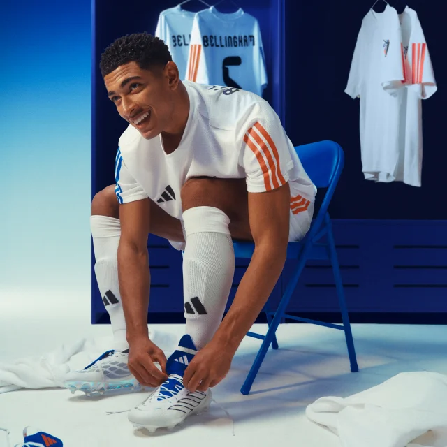 Jude Bellingham ties his red, white, and blue adidas Predator boots while seated in full match kit on a blue chair.