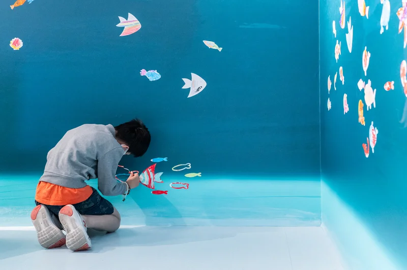 Young visitor kneeling on the floor, adding fish drawings to the wall