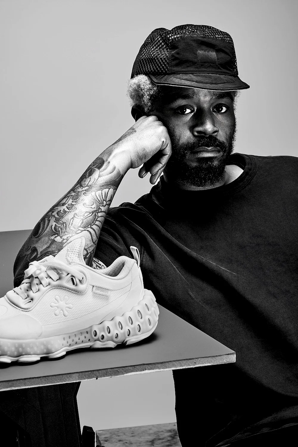 Black-and-white photograph of a person leaning on a table that holds a sneaker