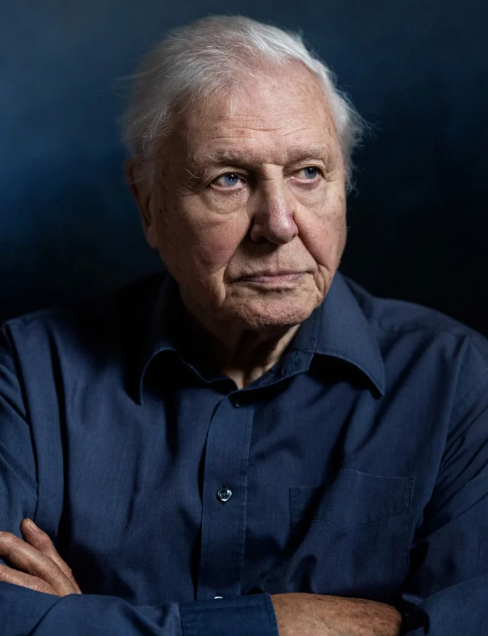 A portrait of Sir David Attenborough with his arms crossed looking to the right.