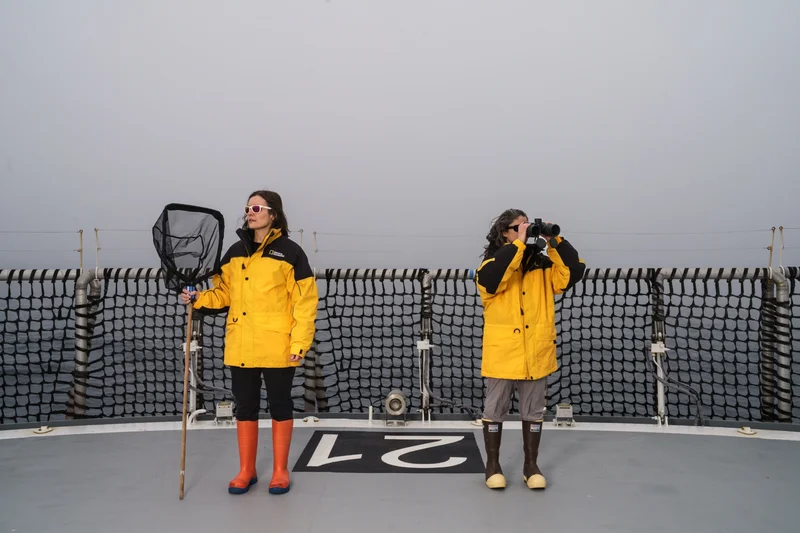 Kim Bernard (left) is an expert on Antarctic krill and is holding a fishing net while wearing a yellow National Geographic jacket. Her colleague Rachel Kaplan (right) is from Oregon State University and is holding up a pair of binoculars while in a yellow National Geographic jacket.
