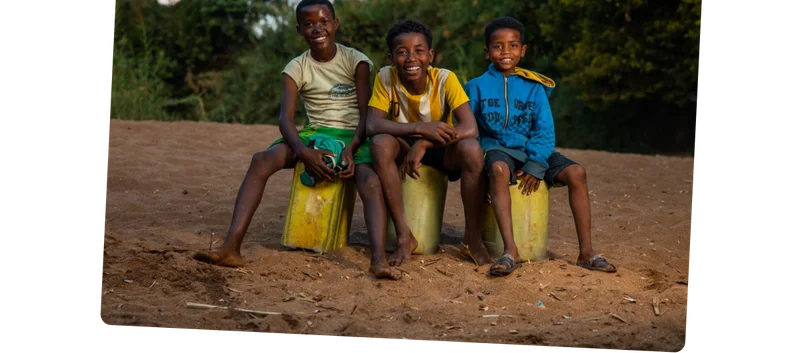 Three young boys sit on top of yellow jerry cans while smiling.