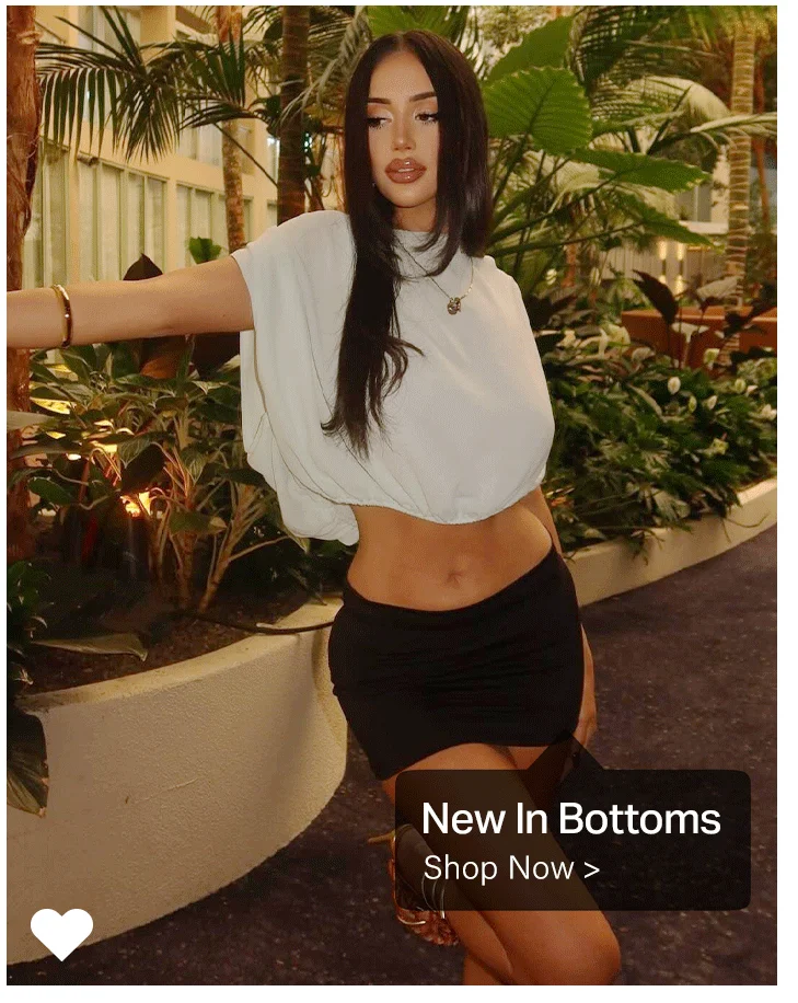 Woman in light grey cropped top and black mini skirt, standing in a lush indoor garden. Discover new arrival bottoms and stylish skirts.