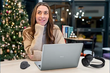 A woman sits at a table behind a Christmas tree with a Dell laptop, mouse and wireless headphones.