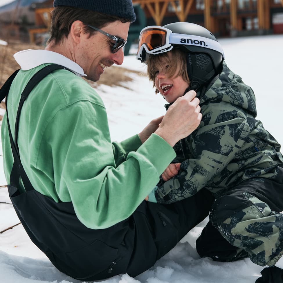 A father and son sitting in the snow wearing Burton outerwear and apparel.
