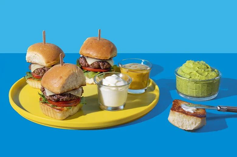 A bright summer-themed food spread featuring three mini burgers, potato chips, pickles, guacamole, and a dipping sauce on a yellow plate and background.