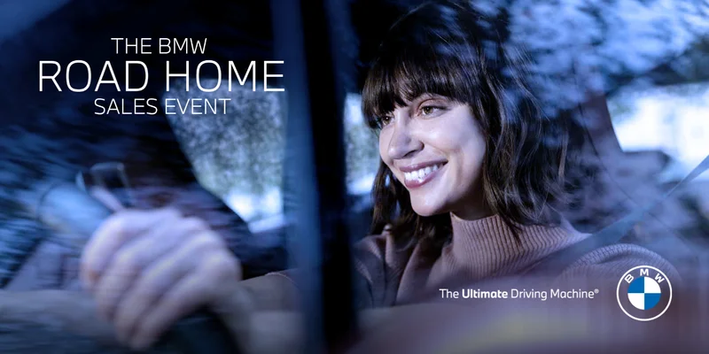 A woman smiling while driving a BMW vehicle. The BMW Road Home Sales Event. The Ultimate Driving Machine®. BMW.