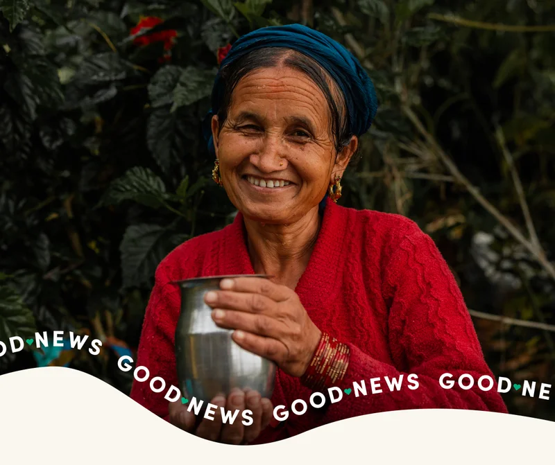 A woman in Nepal holds a cup of clean water with a smile with the Good News logo.