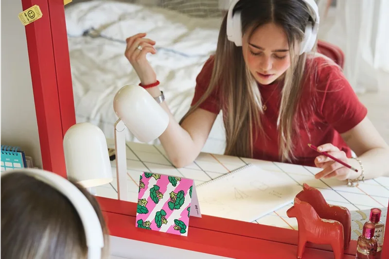A teenager sitting studying by a LAGKAPTEN/SPÄND desk with a colorful grid pattern placed in front of a big red mirror.