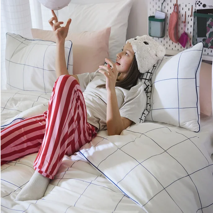 A teenager playing with a round object lying in a bed made with a BREDVECKLARE duvet cover in white with a blue grid pattern.