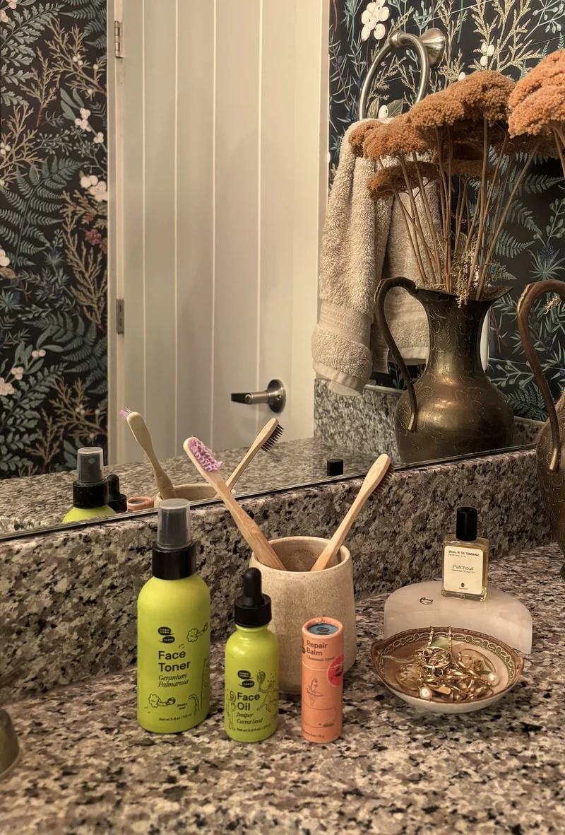 Two green bottles and one orange tube of balm are arranged on a granite counter tip in a bathroom.