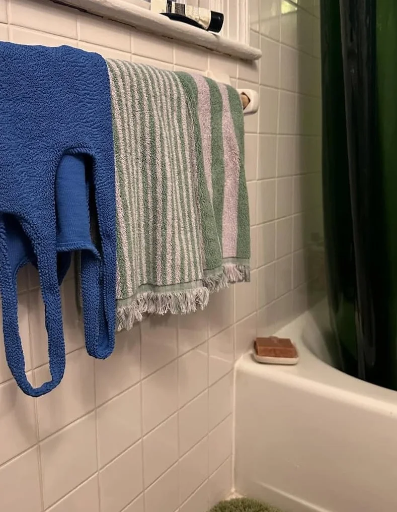 A bar of soap sits on the side of a bathtub with a towel and swimsuit hanging on the towel rack.