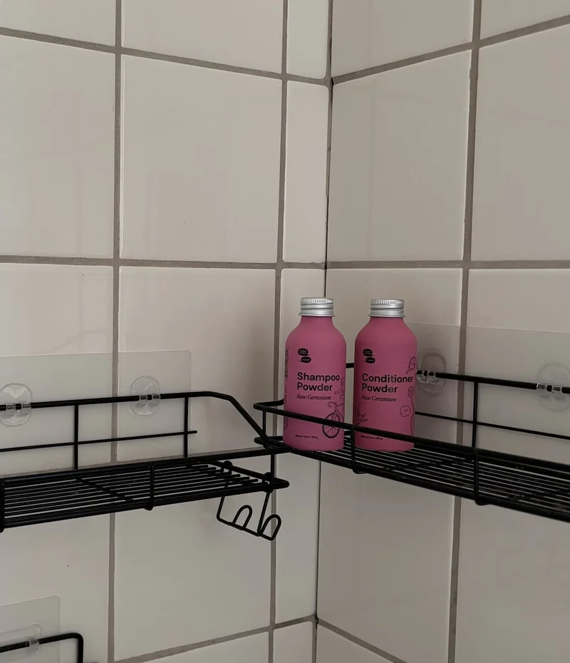 Two pink metal bottles are placed on a black metal rack in a shower with tiled walls.