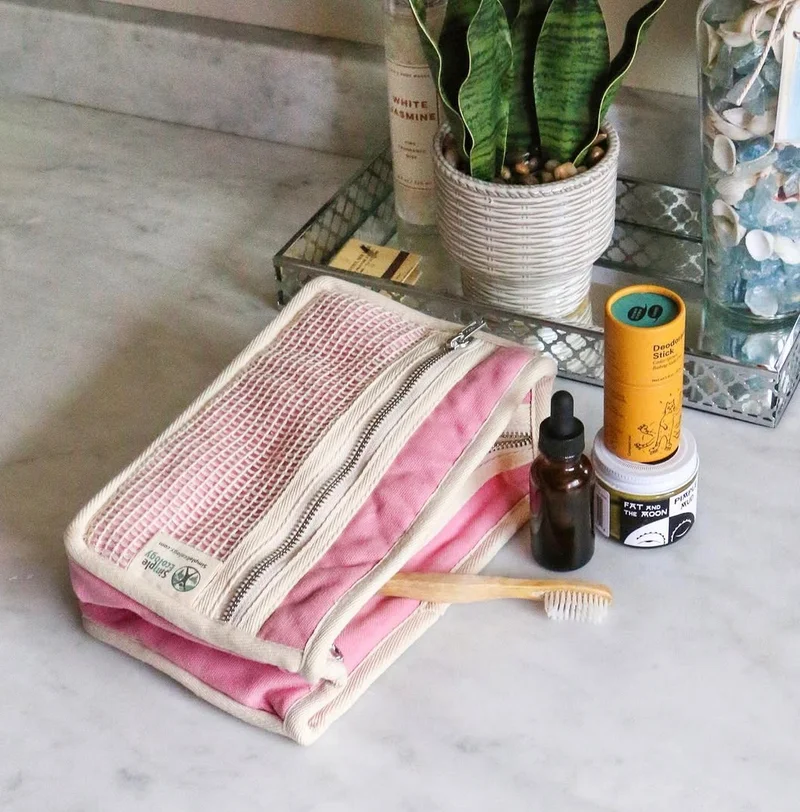 A stick of deodorant in an orange paper tube is arranged next to a few more body care products on a marble counter top with a pink toiletry bag.
