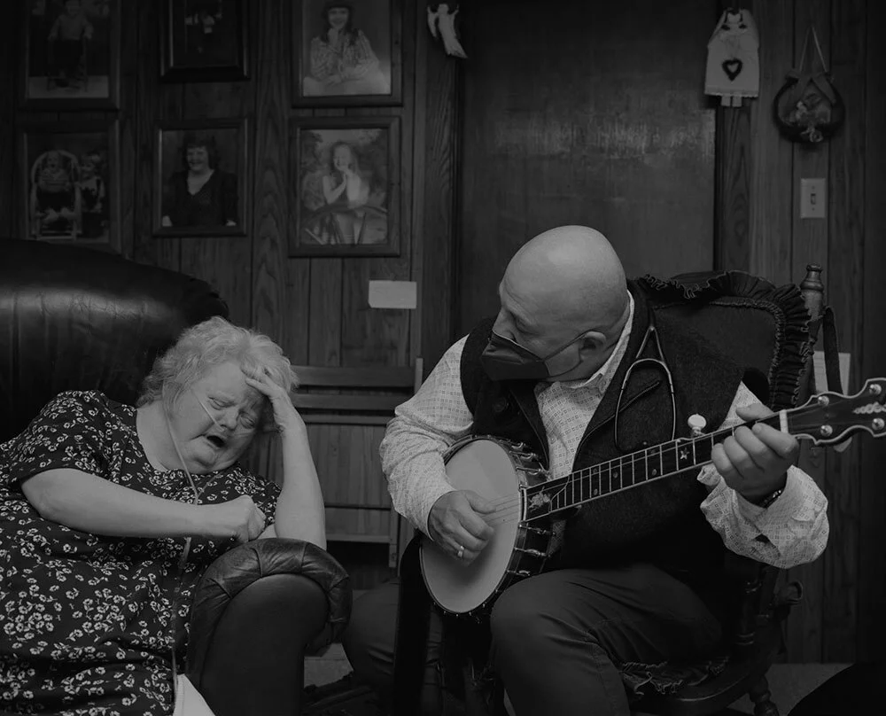 A doctor plays a banjo next to a patient in an armchair