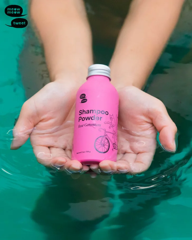 A pair of tan hands holds a pink bottle above a blue water backdrop