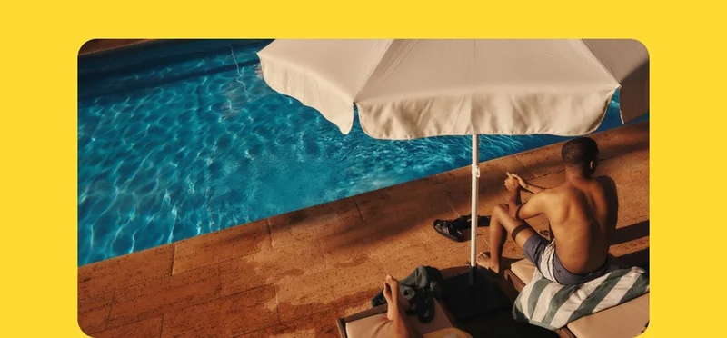 A man in a bathing suit sits poolside under a beige umbrella.