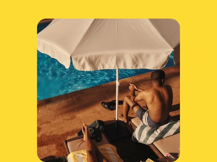 A man in a bathing suit sits poolside under a beige umbrella.