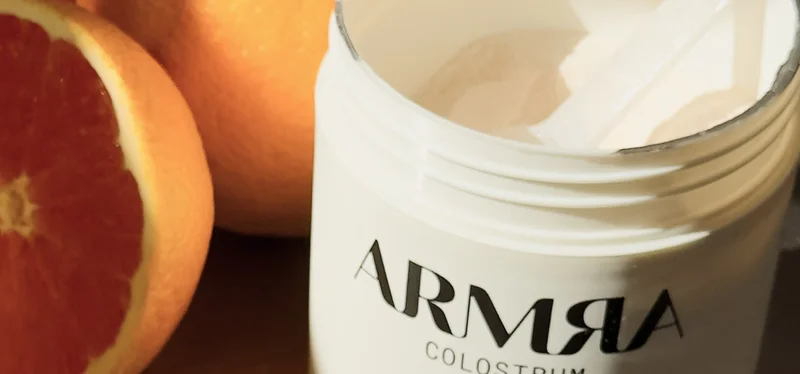Close-up of an open ARMRA Colostrum container with white powder inside, surrounded by fresh halved blood oranges in warm lighting.