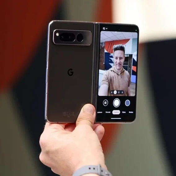 A person’s hand holding a Pixel Fold in one held and taking a selfie. The Pixel Fold shows the person looking into the camera and smiling