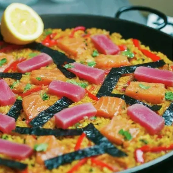A skillet with pieces of salmon and red onion and paella cooking, with a half lemon in the corner.