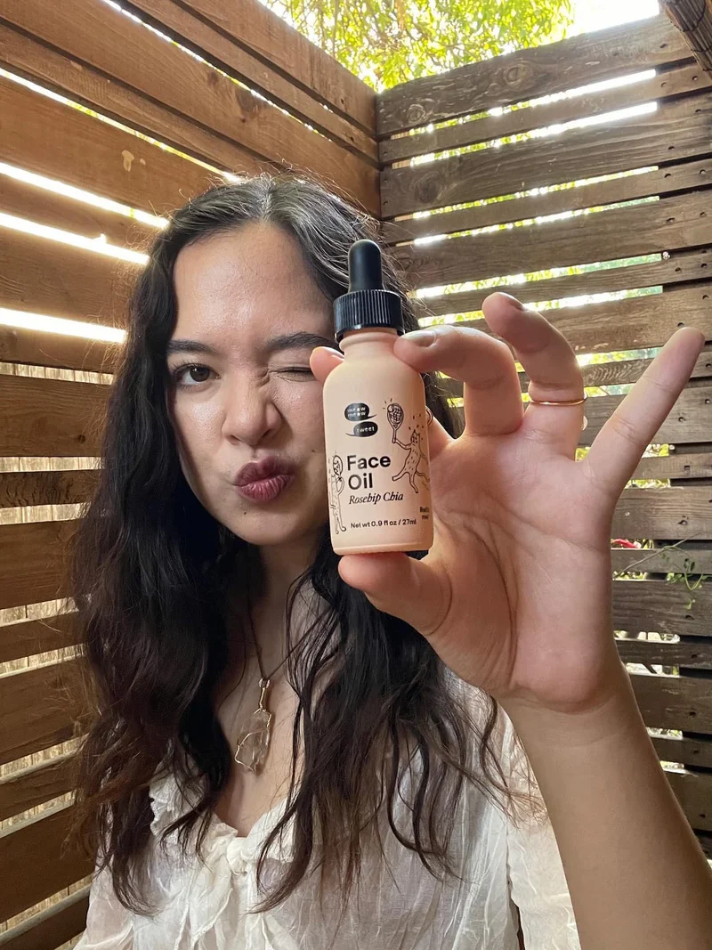 A tan-skinned person holding a face oil bottle up for the camera and smooching her lips in a kiss
