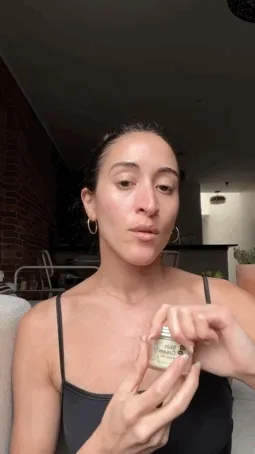 A tan woman with hair in a bun puts skin cream all over her face