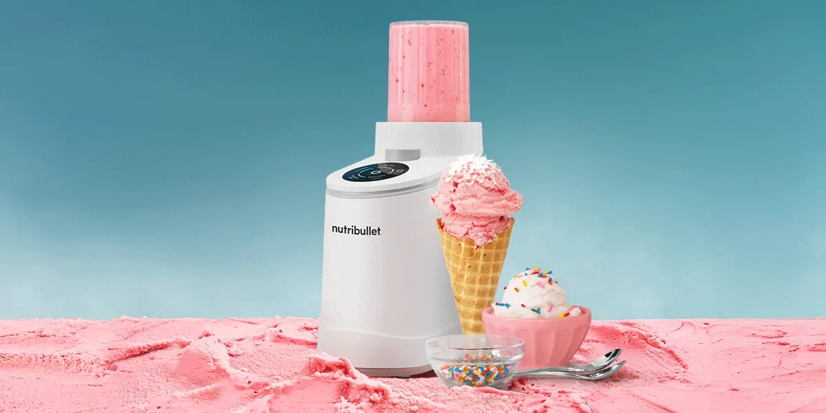 A white nutribullet cream maker sits on pink textured ice cream with a strawberry cone and sprinkle-covered sundae against a gradient blue sky.