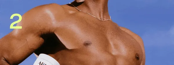 Fit, shirtless person outdoors against a blue sky, flexing their arm while holding a white ARMRA colostrum container.