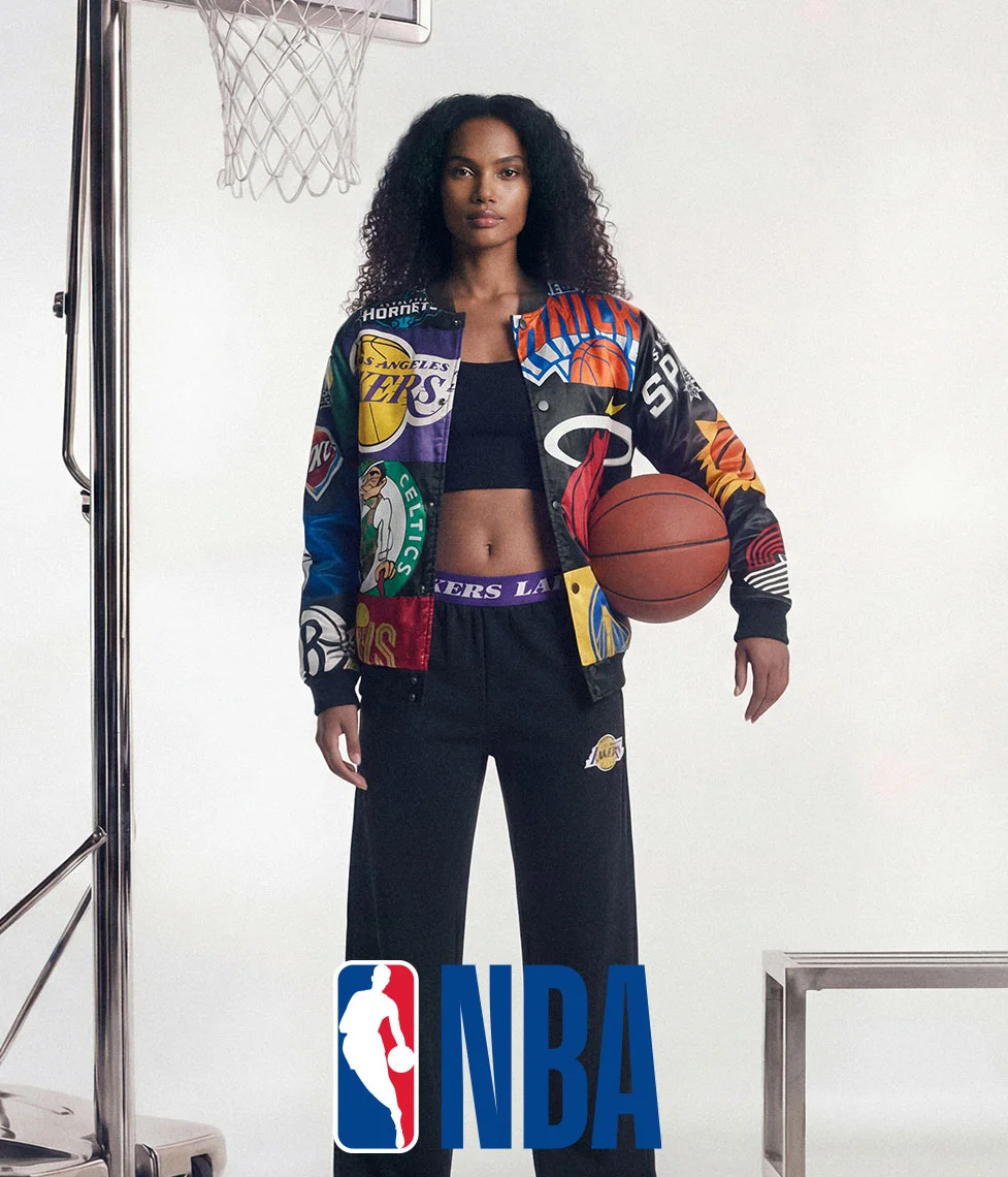 Woman in a vibrant NBA team patch bomber jacket and black athletic pants, holding a basketball next to a hoop. Showcases dynamic NBA fashion.